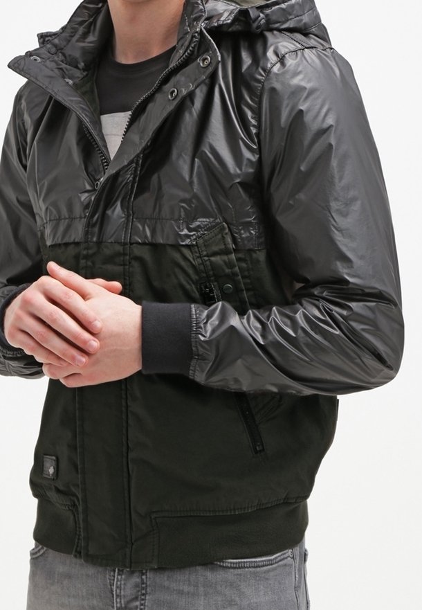 Man i en svart och mörkgrön huva-jacka med glänsande ärmar, stående med händerna knäppta framför sig.