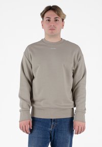 Felpa beige con colletto a girocollo e polsini a costine. Presenta un piccolo logo "Calvin Klein" sul petto. Indossata con jeans in denim blu.