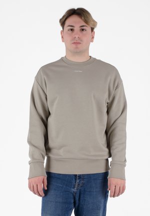 Felpa beige con colletto a girocollo e polsini a costine. Presenta un piccolo logo "Calvin Klein" sul petto. Indossata con jeans in denim blu.