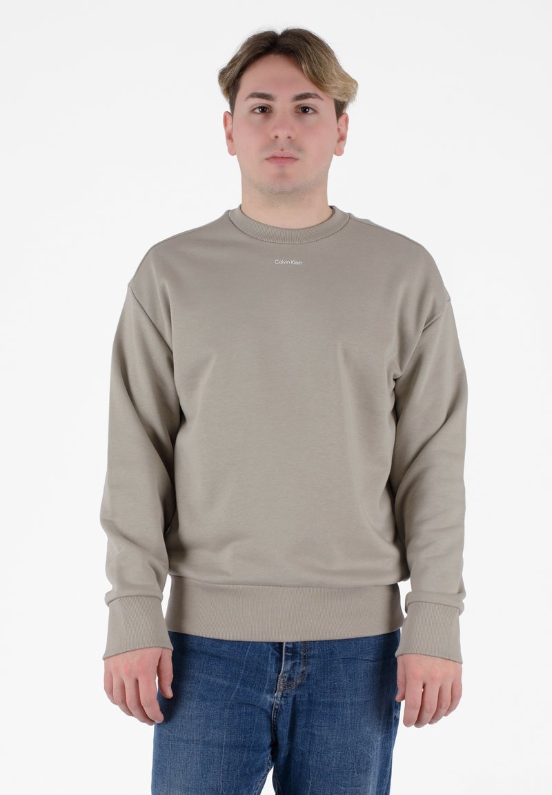 Felpa beige con colletto a girocollo e polsini a costine. Presenta un piccolo logo "Calvin Klein" sul petto. Indossata con jeans in denim blu.