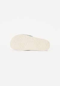 Gummisåle med sikksakkmønster, i en lys beige farge. Overflaten er glatt med en svak glans. Utsmykket med en logo.