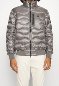 Giubbotto imbottito grigio con colletto alto, design trapuntato a onde, chiusura frontale con zip, tasca con zip nera sul petto e polsini a costina.