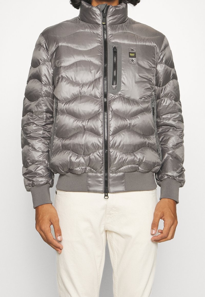 Giubbotto imbottito grigio con colletto alto, design trapuntato a onde, chiusura frontale con zip, tasca con zip nera sul petto e polsini a costina.