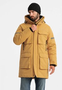 Geel geïsoleerde parka met een capuchon, meerdere voorzakken, ritssluitingen en een grijze accentstreep op de arm. Draag het over een zwarte trui.