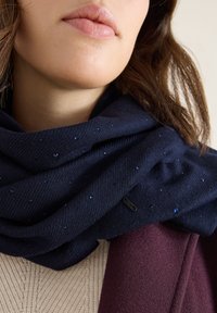 Écharpe navy en tissu doux avec des motifs texturés et de petits accents bleus. Portée par-dessus un vêtement tricoté beige clair et un manteau bordeaux.