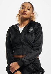 Juoda trumpa zip-up striukė, pagaminta iš minkštos medžiagos, su subtiliu logotipu ant krūtinės, elastiniais rankogaliais ir atitinkamomis kelnėmis. Patogus prigludimas.