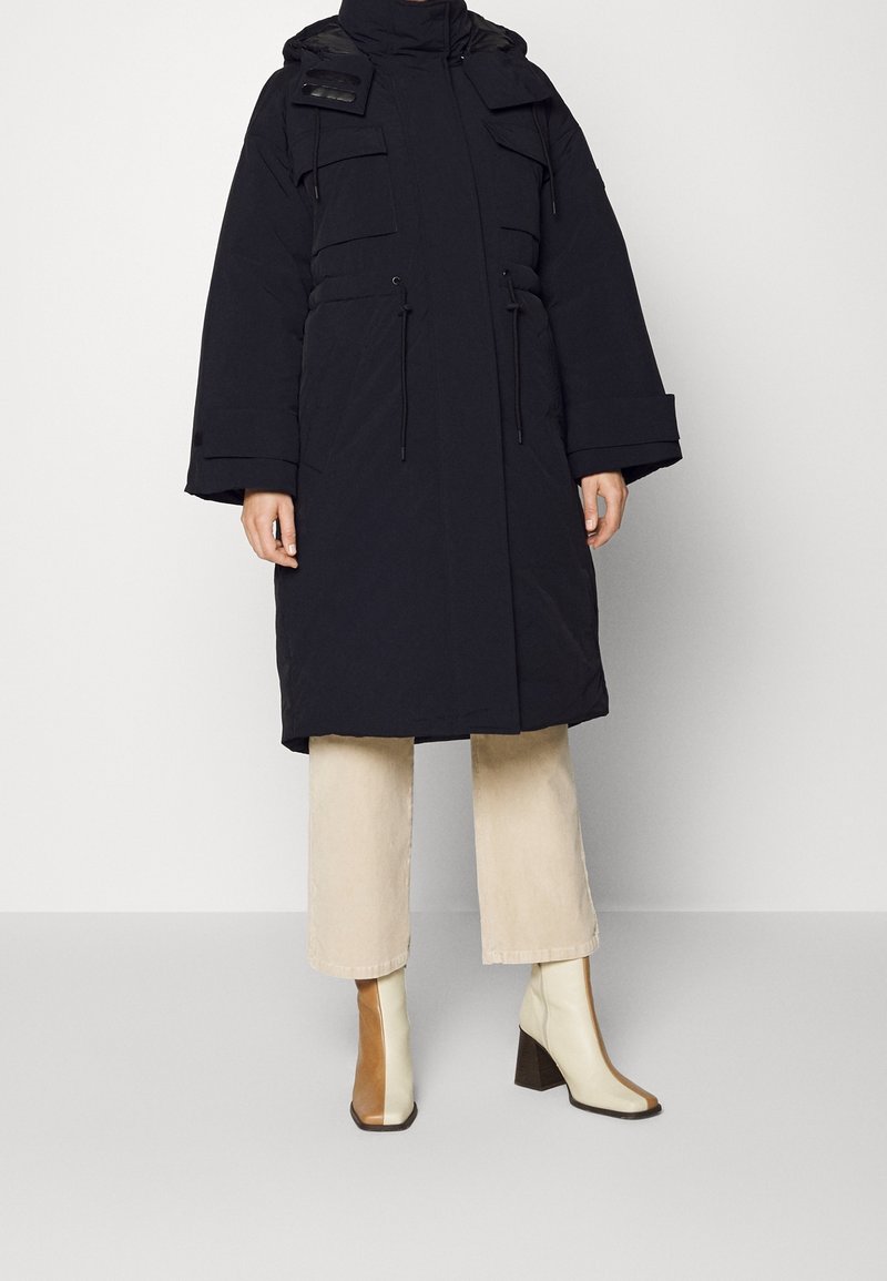 Cappotto imbottito nero con vestibilità oversize, grandi tasche, vita regolabile con coulisse e colletto alto. Abbinato a pantaloni beige a gamba larga e stivali bicolore.