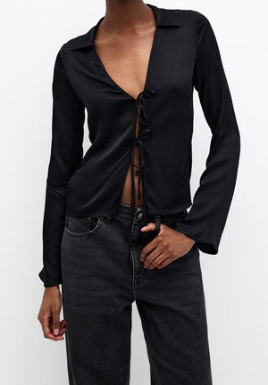 Woman wearing a black long-sleeve tie-front top with deep neckline and high-waisted black jeans, hand resting near pocket.