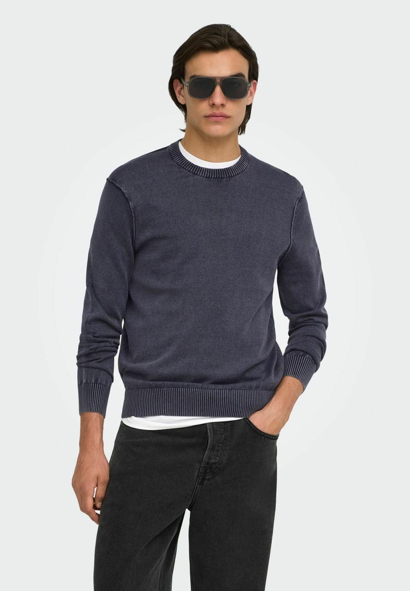 Homme portant un pull bleu foncé, un sous-vêtement blanc, un jean noir et des lunettes de soleil noires, avec une main dans la poche sur fond blanc.