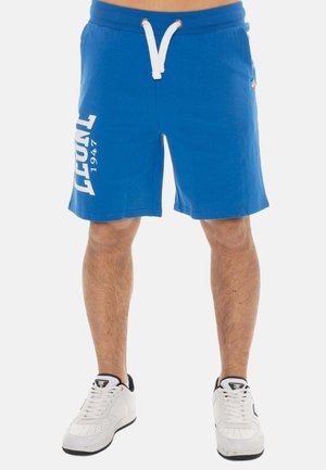 Homme portant un short bleu avec un cordon blanc et des baskets blanches, texte "LOTT 1947" imprimé verticalement sur la jambe gauche.
