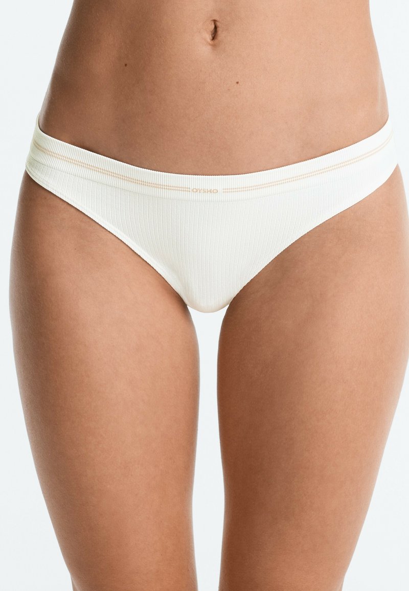 Personne portant un sous-vêtement blanc côtelé style bikini, milieu du corps et haut des cuisses visibles, fond neutre.