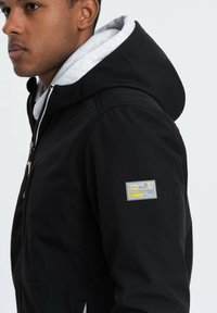 Giacca nera con zip e cappuccio. Dotata di una fodera interna grigia, due tasche con zip e una toppa quadrata con logo sulla manica sinistra.