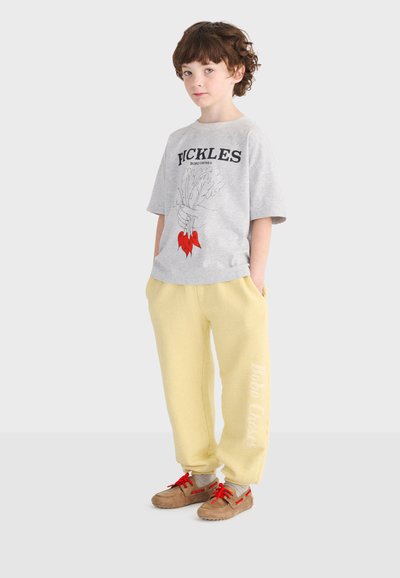 Kind mit lockigem Haar, das ein graues T-Shirt mit Grafik, hellgelbe Jogginghosen mit Text am Bein und braune Schuhe mit roten Schnürsenkeln trägt.