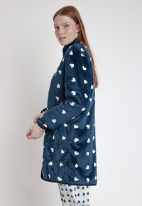 Albornoz de forro polar en azul marino con patrones de corazones blancos. Cuenta con mangas largas y bolsillos laterales, textura suave y ajuste relajado.