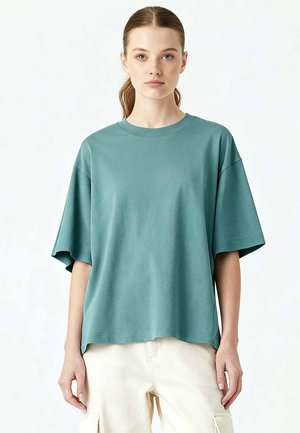 Jeune femme aux cheveux châtain clair attachés en queue de cheval lâche, portant un t-shirt turquoise surdimensionné à manches courtes et un pantalon cargo crème, se tenant face à l'avant.