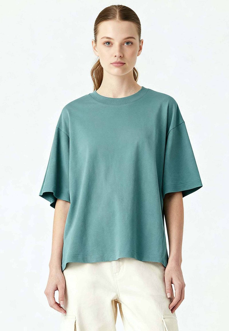 Jeune femme aux cheveux châtain clair attachés en queue de cheval lâche, portant un t-shirt turquoise surdimensionné à manches courtes et un pantalon cargo crème, se tenant face à l'avant.