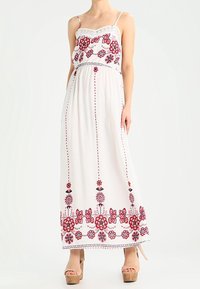 Maxiklänning i vit tyg med röd blommig broderi och vertikala linjer. Har tunna axelremmar och en samlad midja.
