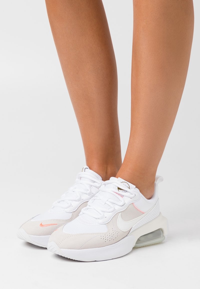 Baskets Nike blanches et beige avec des accents orange clair portées aux pieds sur un fond blanc uni.