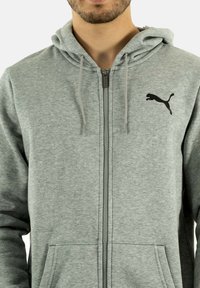 Gray zip-up hoodie made of soft fabric, featuring a black Puma logo on the left chest, drawstring hood, and front pocket.