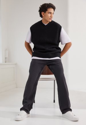 Svart ermeløs strikkegenser over en hvit skjorte, kombinert med mørke jeans og hvite sneakers. Modell sitter på en stol mot en lys bakgrunn.