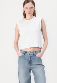 Hvit, ermeløs cropped genser med en teksturert strikkemønster og ribbestrikkede kanter, kombinert med avslappede lyseblå denimjeans.