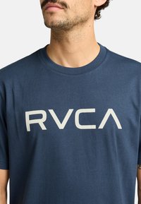 Camiseta de algodón azul marino con un gran logo blanco de RVCA en el frente. Mangas cortas y escote redondo. Textura suave.