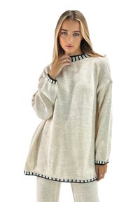 Suéter de punto oversize beige con cuello y puños a rayas negras, que presenta un acabado texturizado y un distintivo detalle de flecos en el dobladillo.