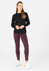 Langarm schwarzes Oberteil kombiniert mit weinroten Leggings, die eine glatte Textur aufweisen. Weiße Sportschuhe mit orangefarbenen und türkisen Akzenten.