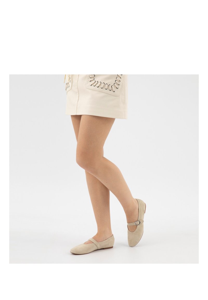 Femme portant des ballerines Mary Jane en suède beige et une mini-jupe crème avec des détails de poches cousues, debout jambes croisées sur fond blanc.