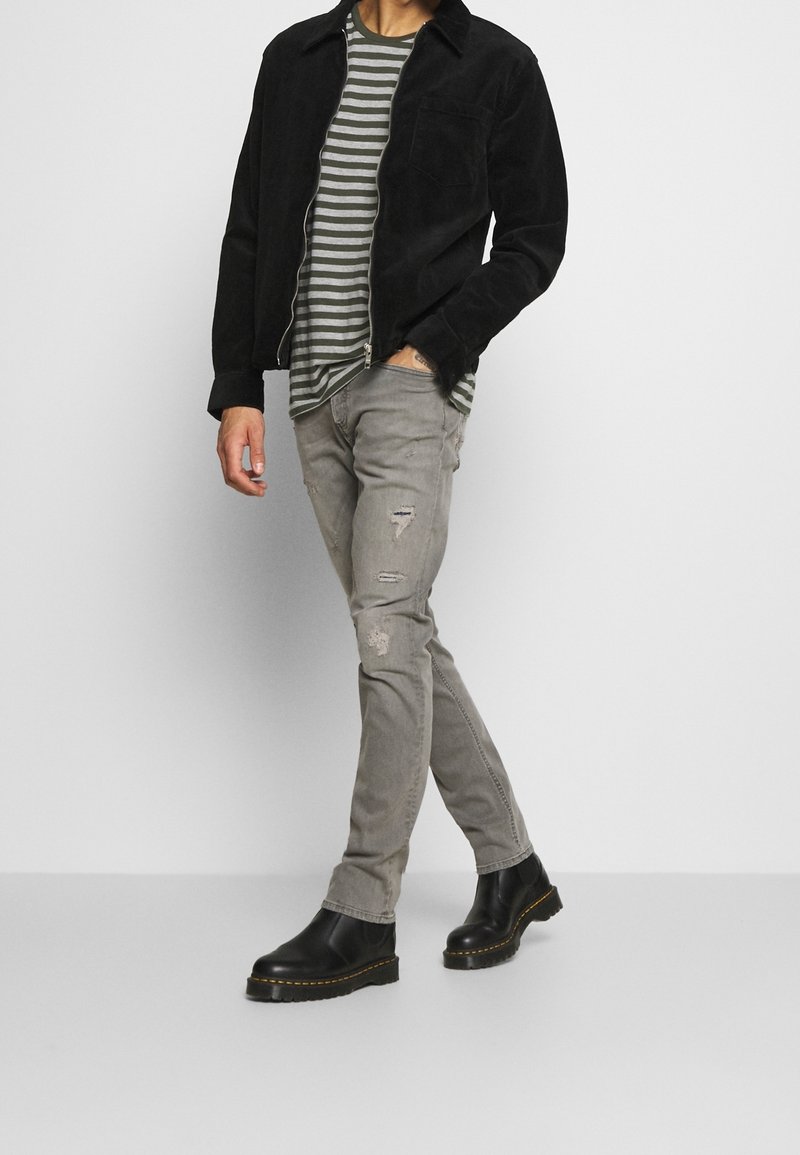 Homme portant une veste noire, une chemise rayée grise, un jean gris délavé et des bottes noires, debout devant un fond clair uni.