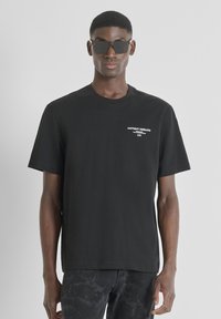 Camiseta de algodón negra con mangas cortas y cuello redondo. Presenta un logotipo impreso en blanco en el pecho izquierdo. El modelo lleva gafas de sol negras en forma rectangular.