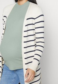 Ribbade koftan i grädde med marinblå ränder, parat med en enfärgad grön topp. Har långa ärmar och en avslappnad passform. Denimjeans synliga.