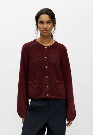 Femme aux cheveux foncés portant un cardigan bordeaux boutonné avec deux poches avant et un pantalon noir, debout devant un fond clair uni.