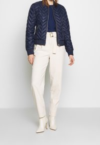 Námořnická prošívaná bomber bunda s chevronovým vzorem, v kombinaci s krémovými kalhotami s vysokým pasem a bílými botami na podpatku do kotníku.