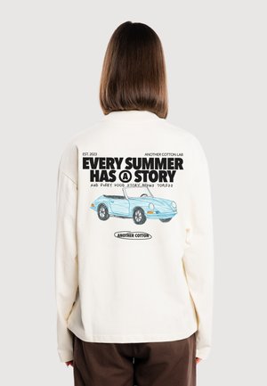 Persona que lleva una camiseta de manga larga color crema con texto negro y gráfico de un coche descapotable azul en la espalda que dice "Cada verano tiene una historia."