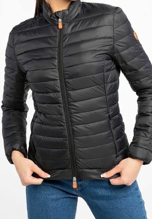 Czarna pikowana kurtka z przednim zamkiem błyskawicznym i pomarańczowymi suwakami, noszona przez osobę poprawiającą dół kurtki, w połączeniu z niebieskimi dżinsami.