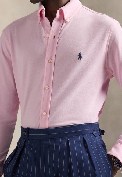 Light pink button-up shirt with a navy embroidered logo, paired with navy pinstripe trousers featuring a visible waistband and buttons.