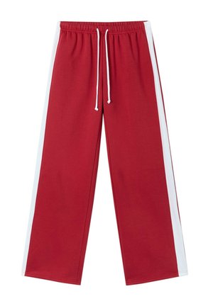 Jeune femme debout en survêtement rouge et blanc avec le numéro 24 sur le sweat à capuche, portant des chaussures blanches sur un fond gris uni.