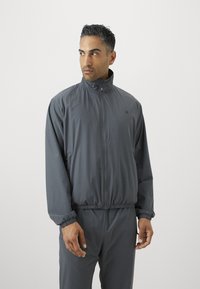 Mann som har på seg en mørkegrå zip-up jakke og matchende bukser, som ser til venstre mot en enkel lys bakgrunn.