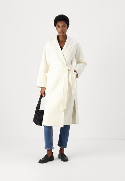 Weißer Wickelmantel mit großem Kragen, Seitentaschen und einem gebundenen Gürtel, kombiniert mit blauen, gekürzten Jeans und schwarzen Slip-on-Schuhen. Schwarze gewebte Tasche.