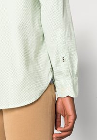 Camicia verde chiaro con un piccolo motivo a pois. Presenta polsini con bottoni e spacco laterale. Indossata con pantaloni beige, che mostrano una superficie di tessuto liscia.