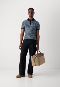 Granatowa koszulka polo z wzorzystym deseniem, krótki rękaw i kołnierz; zestawiona z ciemnoniebieskimi spodniami i beżową torbą podróżną. Jasnobrązowe buty.