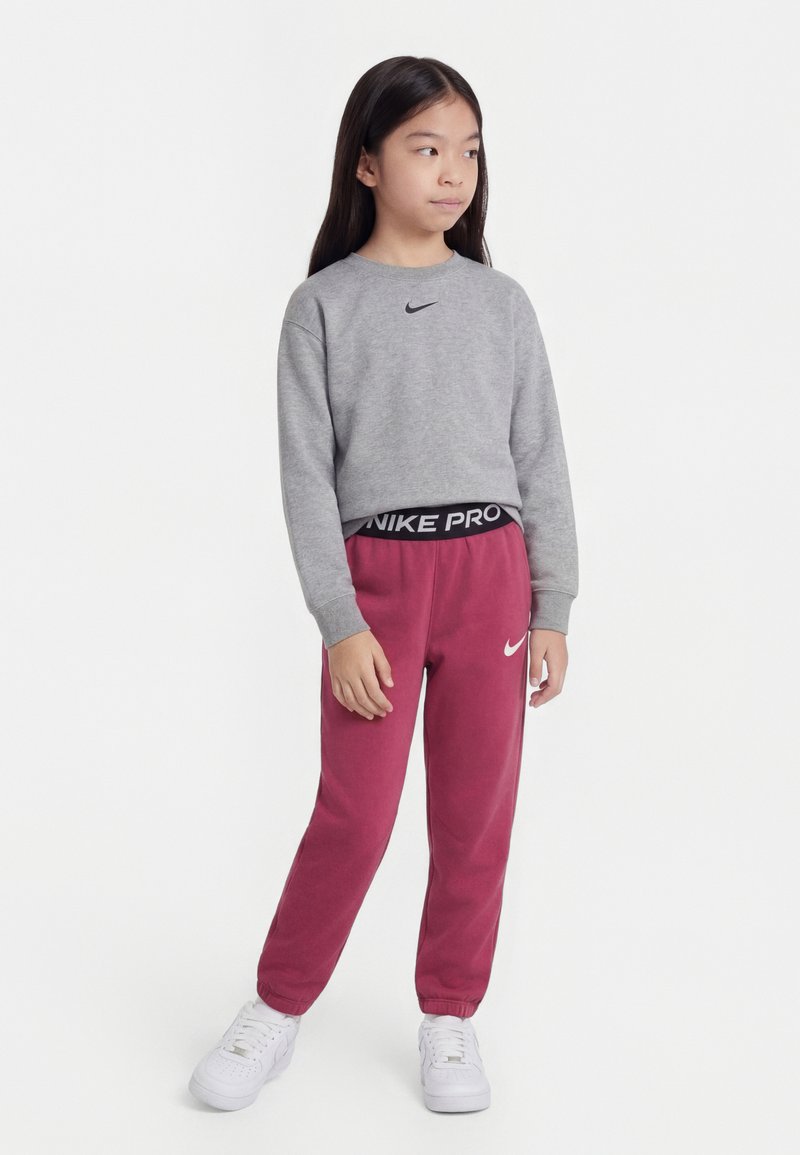 Młoda dziewczyna ubrana w szarą bluzę Nike, bordowe spodnie dresowe Nike Pro i białe buty sportowe, stoi z rękami swobodnie opuszczonymi wzdłuż ciała.