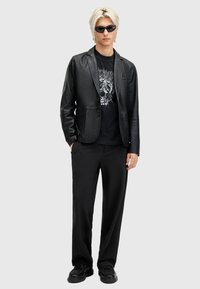 Blazer de cuero negro con solapas, combinado con una camiseta gráfica negra, pantalones holgados negros y zapatos negros, complementado con gafas de sol.