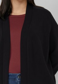 Vrouw draagt een zwarte open cardigan over een bordeauxrood geribbeld topje en een blauwe spijkerbroek met een zichtbare knoopsluiting.