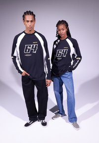 Two models wear matching black long-sleeve shirts with white accents and a large "64" graphic. The man pairs his with black pants; the woman wears denim.