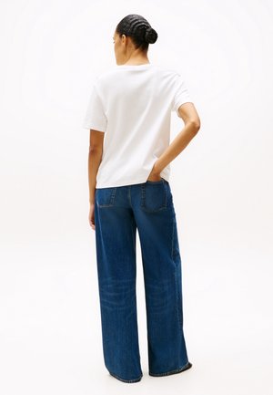 Femme avec les cheveux en chignon portant une chemise blanche à manches courtes et un jean bleu à jambes larges, debout avec une main dans la poche arrière.