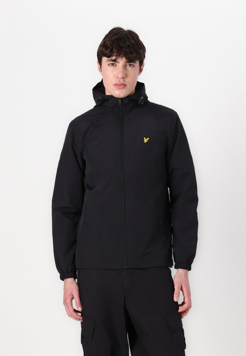 Schwarze Jacke mit einem Reißverschluss vorne, elastischen Bündchen und verstellbare Kapuze. Mit kleinem gelben Logo auf der Brust. Glatte Textur.