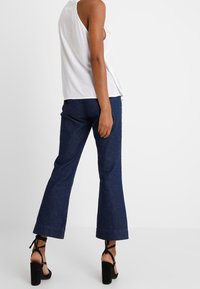 Weißes ärmelloses Oberteil kombiniert mit dunkelblauen Schlagjeans. Das Outfit hat eine lässige Passform und schwarze High Heels mit Knöchelriemen.