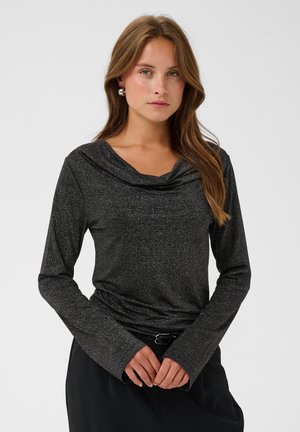 Femme aux longs cheveux bruns portant un haut noir ajusté à manches longues scintillant et un pantalon noir, debout devant un fond blanc uniforme.